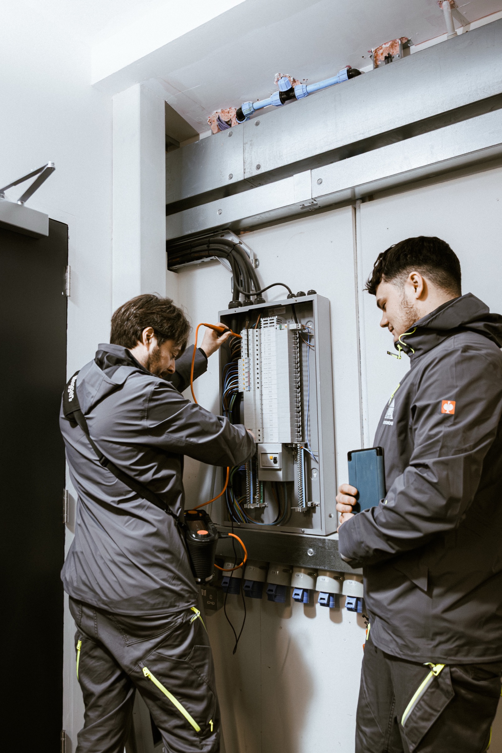 Two Autonomous engineers in branded olive workwear flanking an open electrical distribution board mid-installation.
