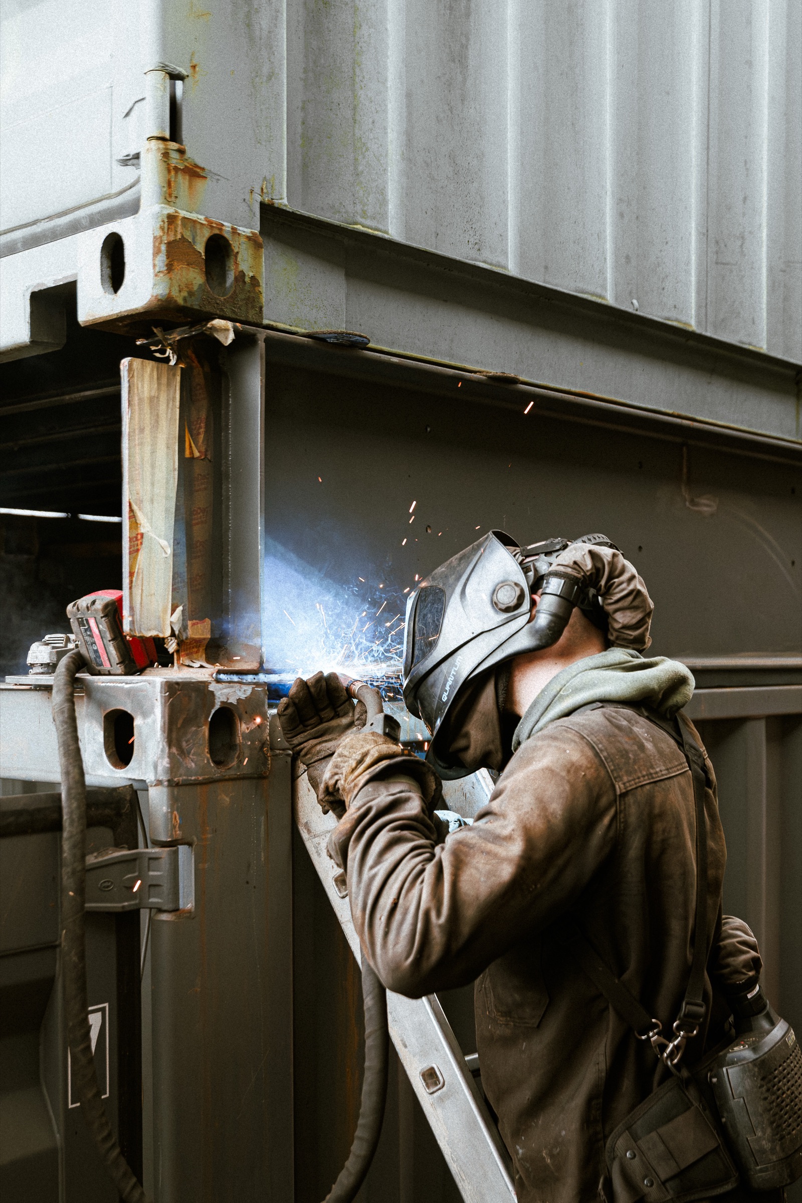 An engineer in heavy canvas workwear MIG-welding on the corner of an ISO container, sparks visible against the corrugated steel.