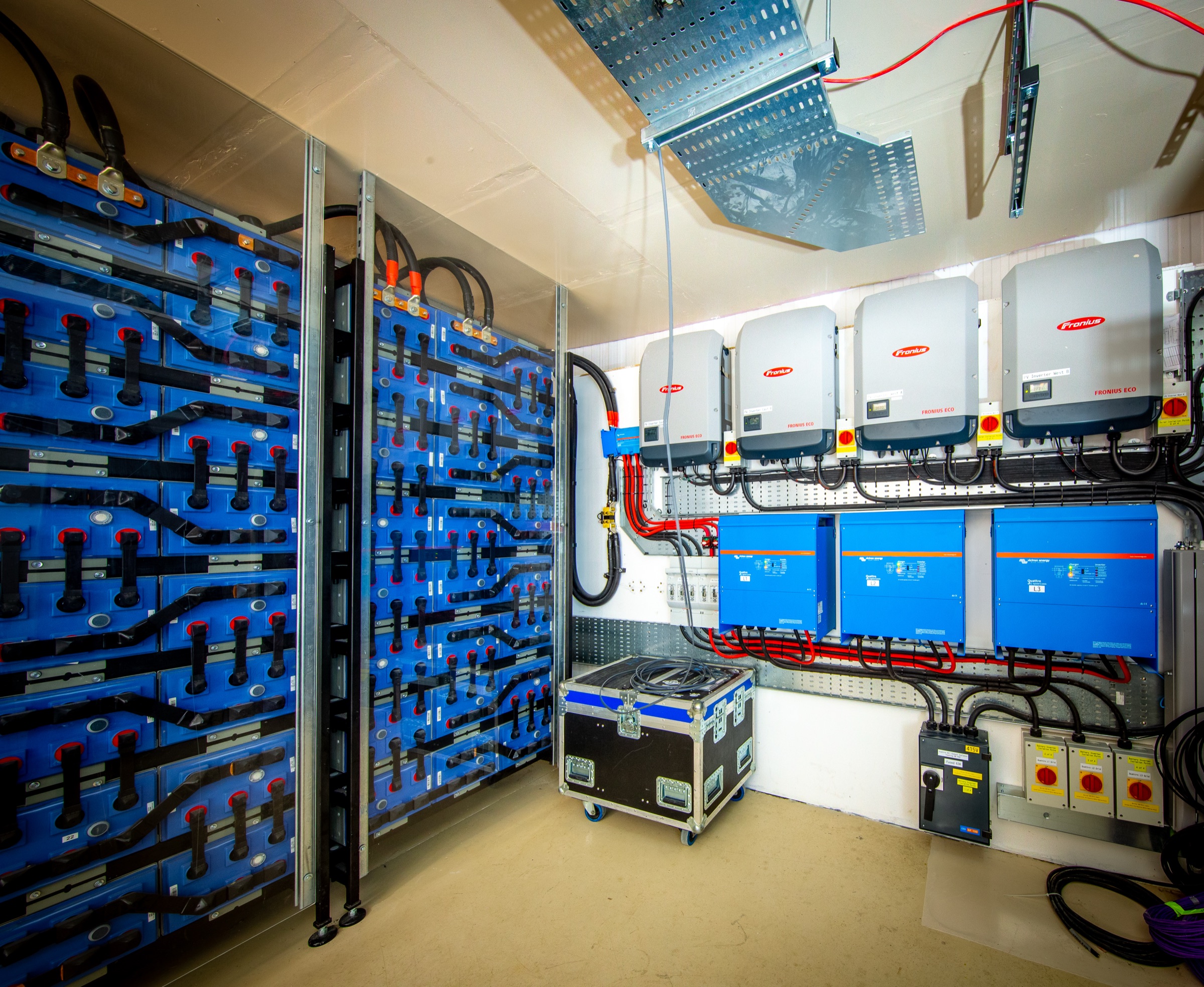 A finished off-grid power station: a stack of OPzS lead-acid traction cells with red caps, four Fronius string inverters and three Victron Quattro inverter/chargers, fully labelled.